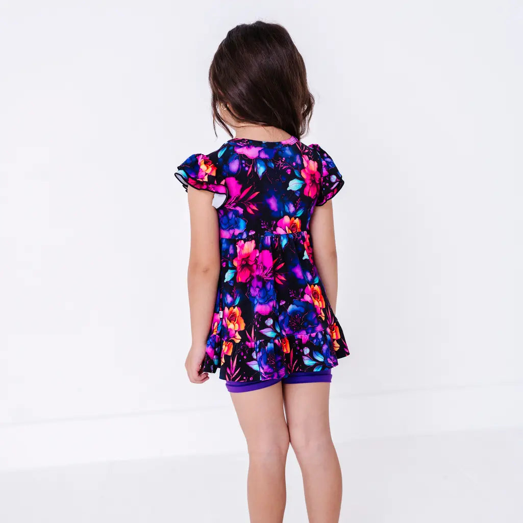 Back view of a child with long hair, in a flutter sleeve peplum top in a black watercolor floral print with purple bike shorts.