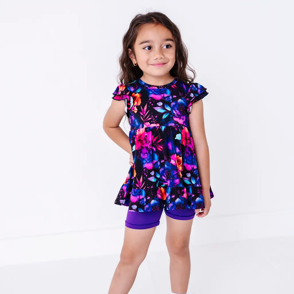 A smiling child with a hand on the hip looking up, in a flutter sleeve peplum top in a black watercolor floral print with purple bike shorts.