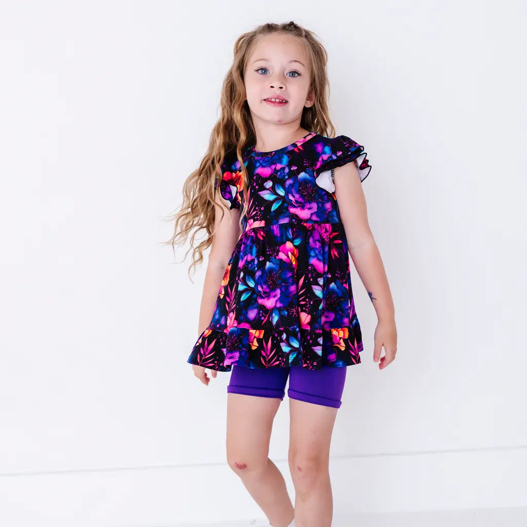A child with long hair standing with the arms at the sides, in a flutter sleeve peplum top in a black watercolor floral print with purple bike shorts.
