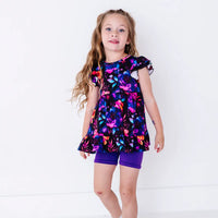 A child with long hair standing with the arms at the sides, in a flutter sleeve peplum top in a black watercolor floral print with purple bike shorts.