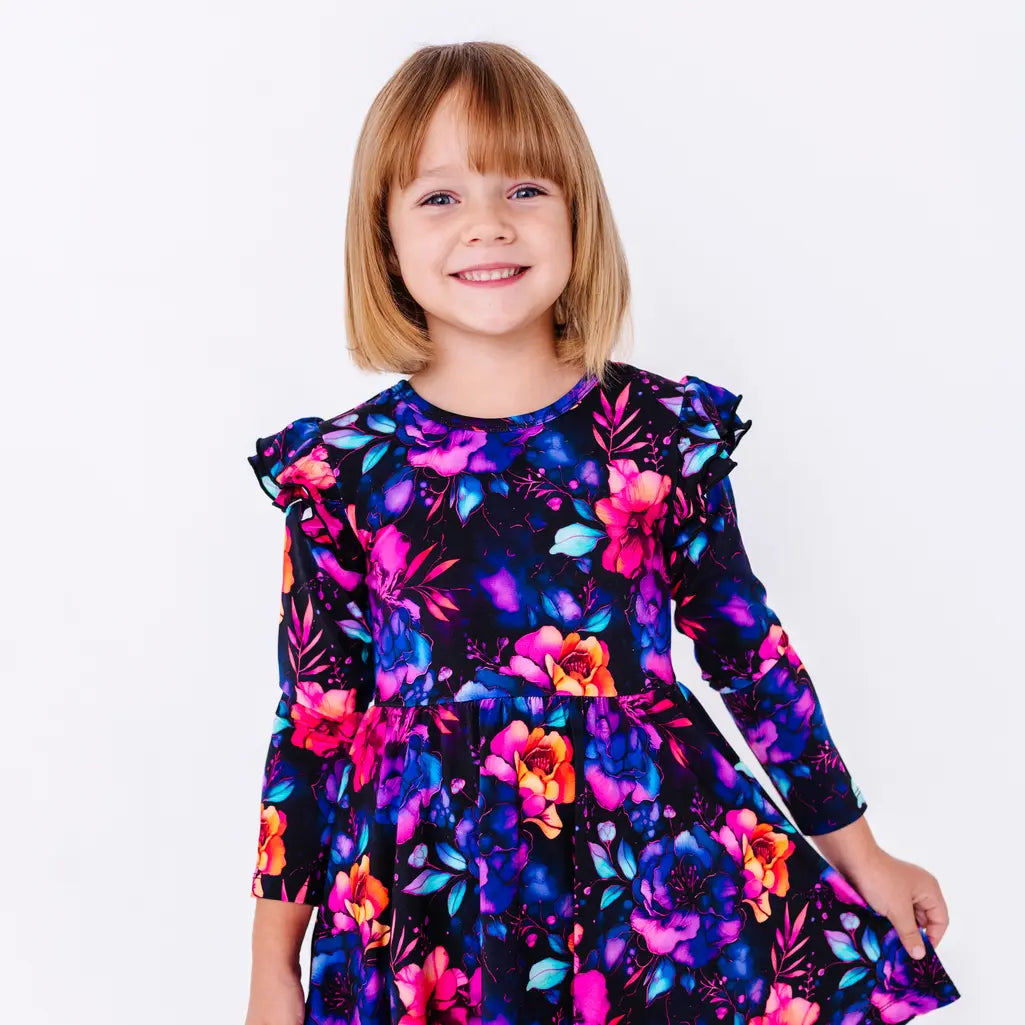 Close up portrait of a child with a big smile, in a long sleeve twirl dress with flutter ruffles at the shoulders in a black print of pink, orange, purple, and blue watercolor flowers.