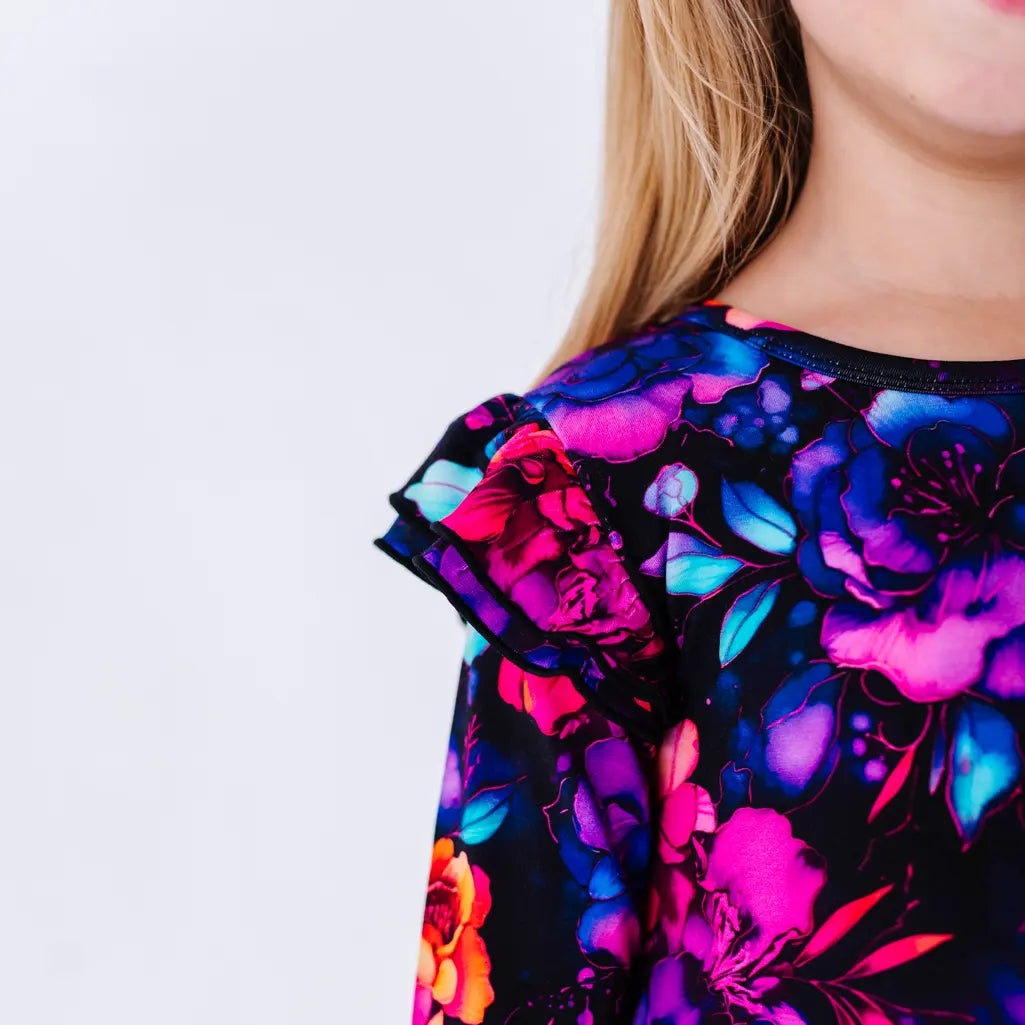 Close up of a flutter ruffle on the shoulder of a long sleeve twirl dress in a black print of pink, orange, purple, and blue watercolor flowers.