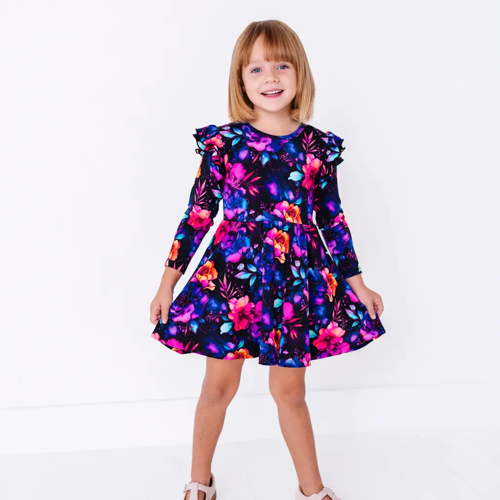 Full length view of a smiling child standing in a long sleeve twirl dress with flutter ruffles at the shoulders in a black print of pink, orange, purple, and blue watercolor flowers.
