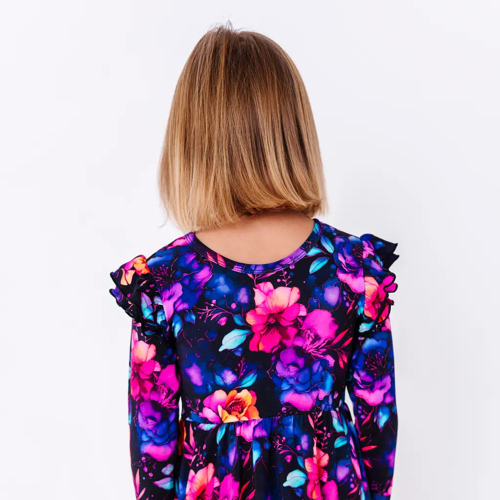 Close up of the back of a child with short hair, in a long sleeve twirl dress with flutter ruffles at the shoulders in a black print of pink, orange, purple, and blue watercolor flowers.