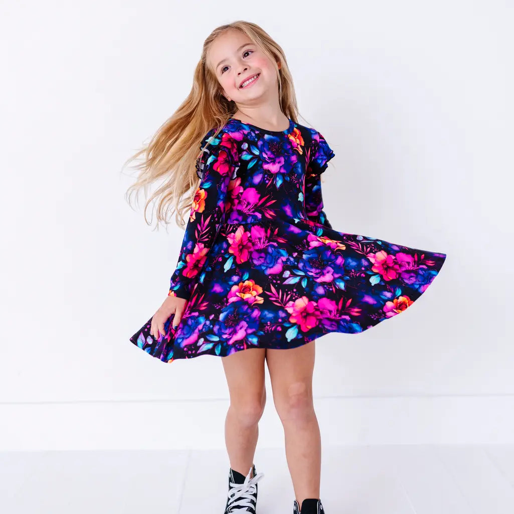 A smiling child twirling with the skirt flared, in a long sleeve twirl dress with flutter ruffles at the shoulders in a black print of pink, orange, purple, and blue watercolor flowers.