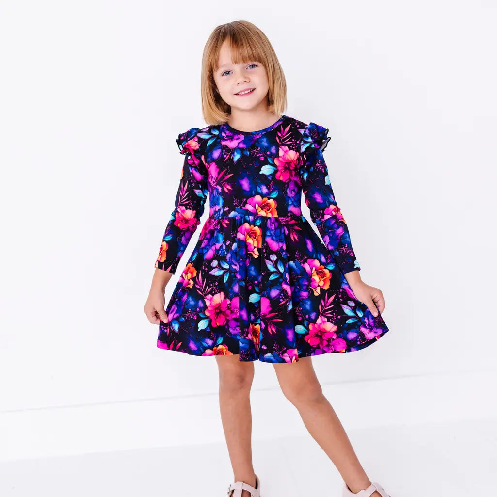A smiling child with short hair holding the skirt, in a long sleeve twirl dress with flutter ruffles at the shoulders in a black print of pink, orange, purple, and blue watercolor flowers.