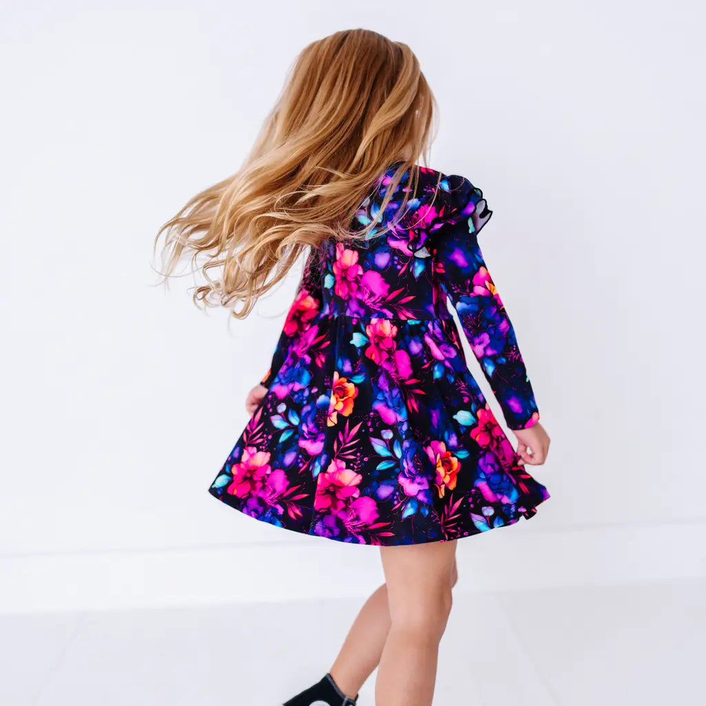Back view of a child twirling with hair flying, in a long sleeve twirl dress with flutter ruffles at the shoulders in a black print of pink, orange, purple, and blue watercolor flowers.
