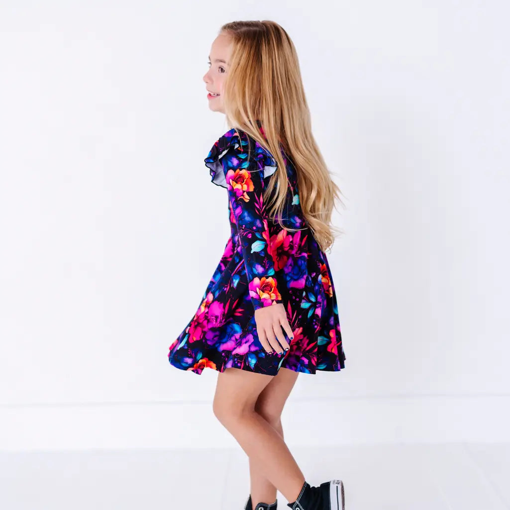 Side view of a child with long hair mid twirl, in a long sleeve twirl dress with flutter ruffles at the shoulders in a black print of pink, orange, purple, and blue watercolor flowers.