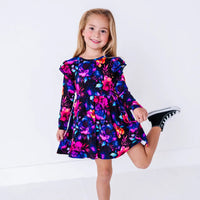 A smiling child with one leg lifted playfully, in a long sleeve twirl dress with flutter ruffles at the shoulders in a black print of pink, orange, purple, and blue watercolor flowers.
