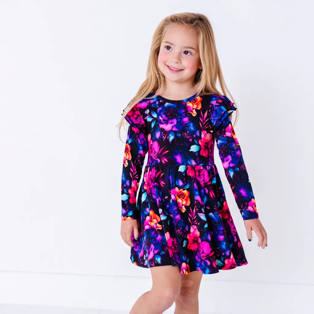 A smiling child walking toward the camera, in a long sleeve twirl dress with flutter ruffles at the shoulders in a black print of pink, orange, purple, and blue watercolor flowers.