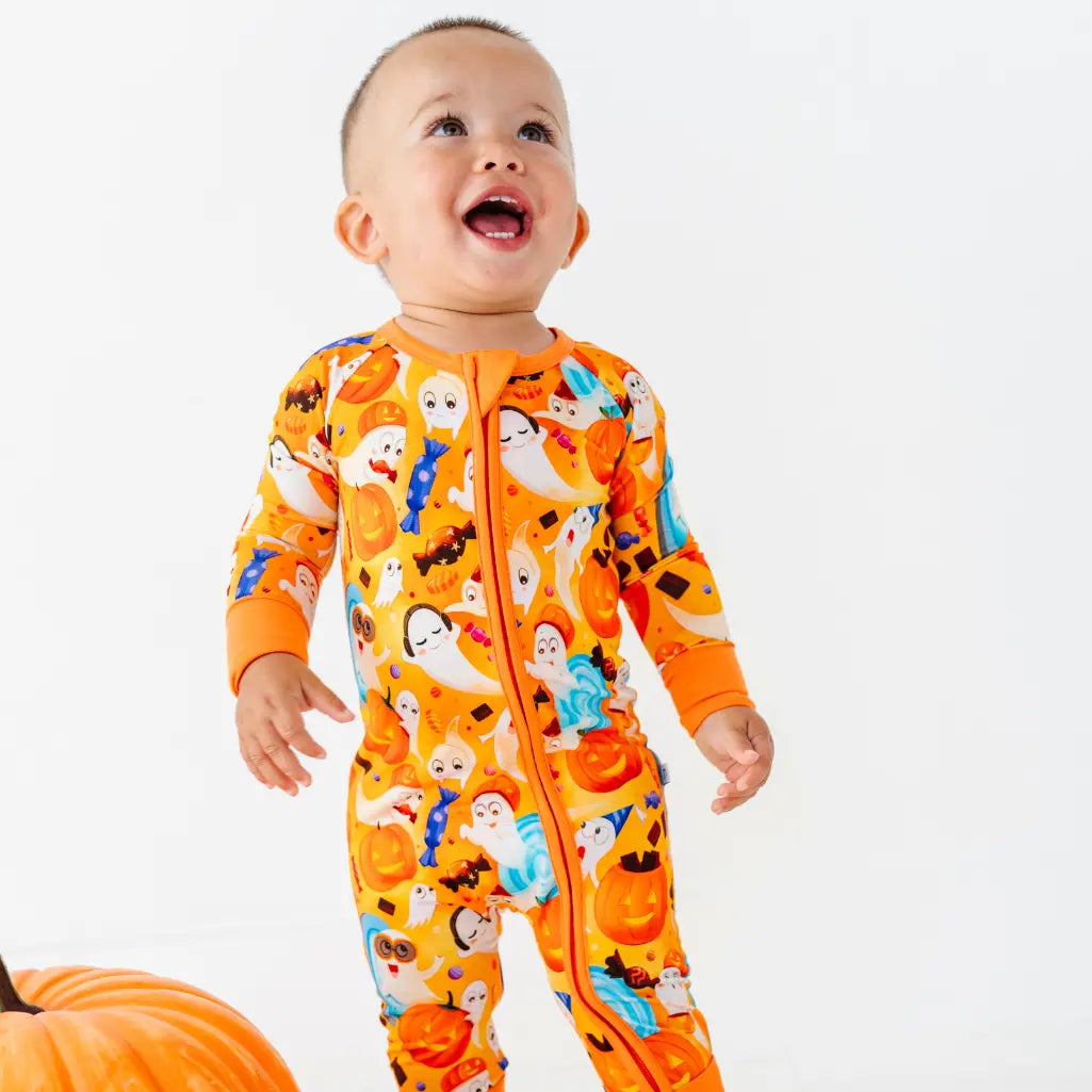 A baby standing and laughing while wearing an orange zip-up pajama with a friendly ghost and pumpkin pattern.