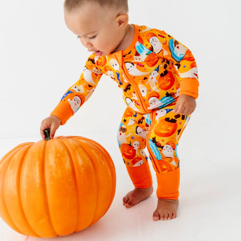 A baby in an orange ghost and pumpkin print zip-up pajama reaching for the stem of a large pumpkin.