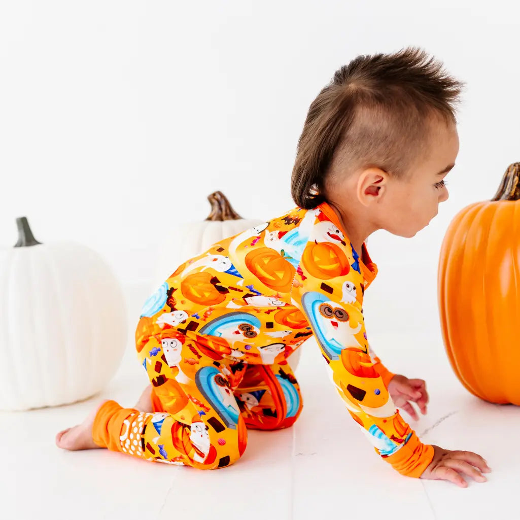 A child crawling between white and orange pumpkins wearing an orange zip-up pajama with a ghost and pumpkin print.