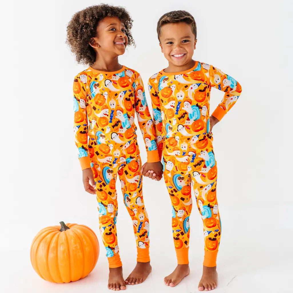 Two children holding hands next to a pumpkin while wearing matching orange two-piece pajamas with a ghost and pumpkin print.