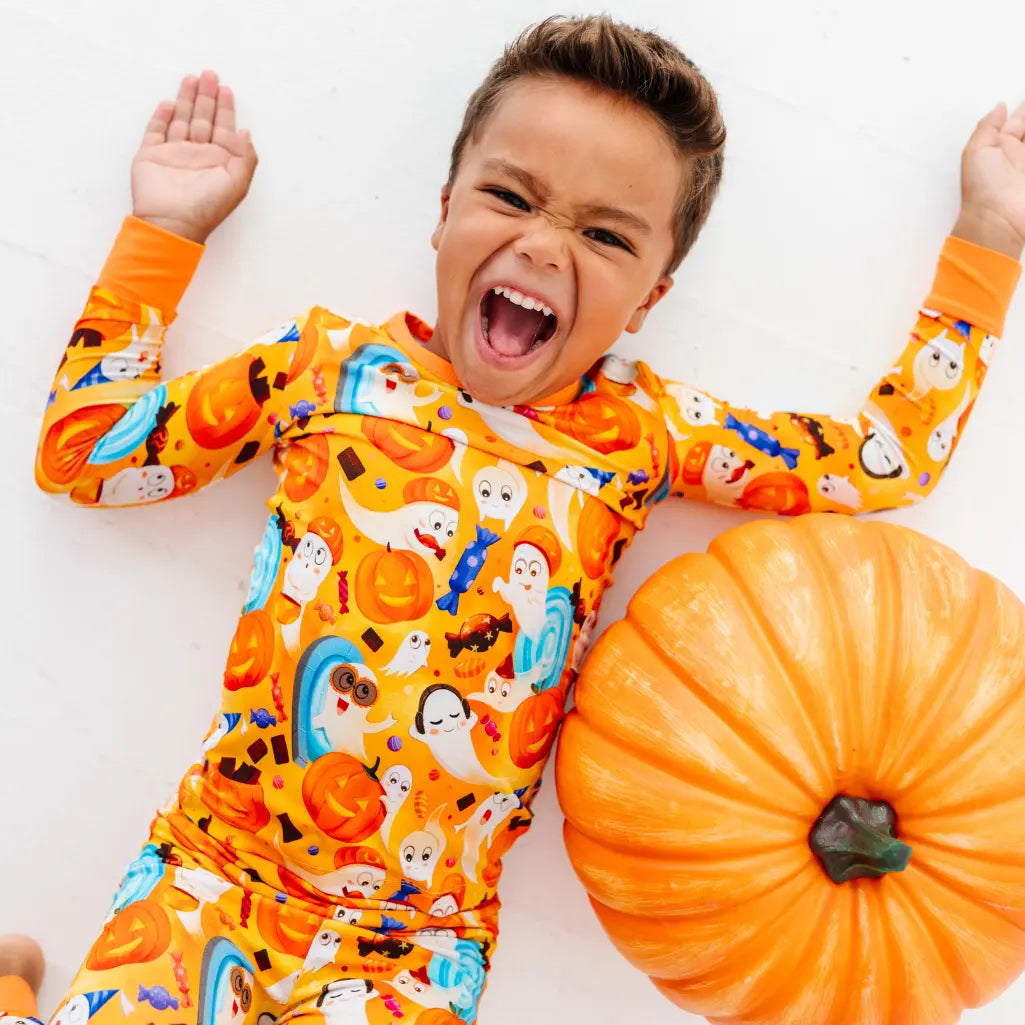 A child lying on a white floor next to a pumpkin, laughing, in an orange pajama set with a friendly ghost print.