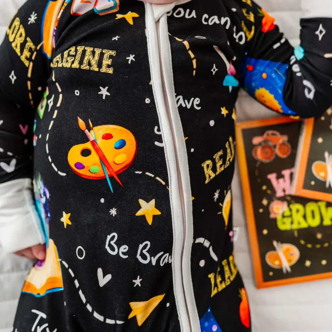 Close-up of a black zip-up baby pajama printed with paint palettes, stars, and words like Imagine, Be Brave, and Learn.