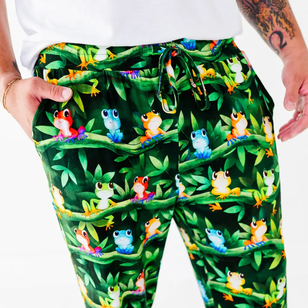 Close-up of a man wearing dark green pajama pants covered in colorful tree frogs on leafy vines with a drawstring waist.