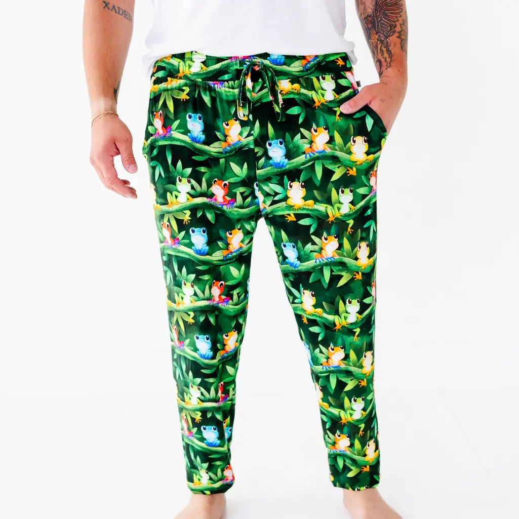 A man standing with one hand in the pocket of dark green pajama pants with a colorful tree frog and jungle leaf print.