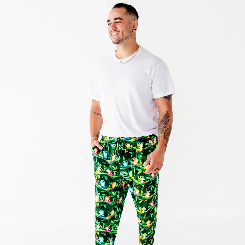 A man in a white tee smiling with his hand in the pocket of dark green pajama pants with a colorful tree frog print.