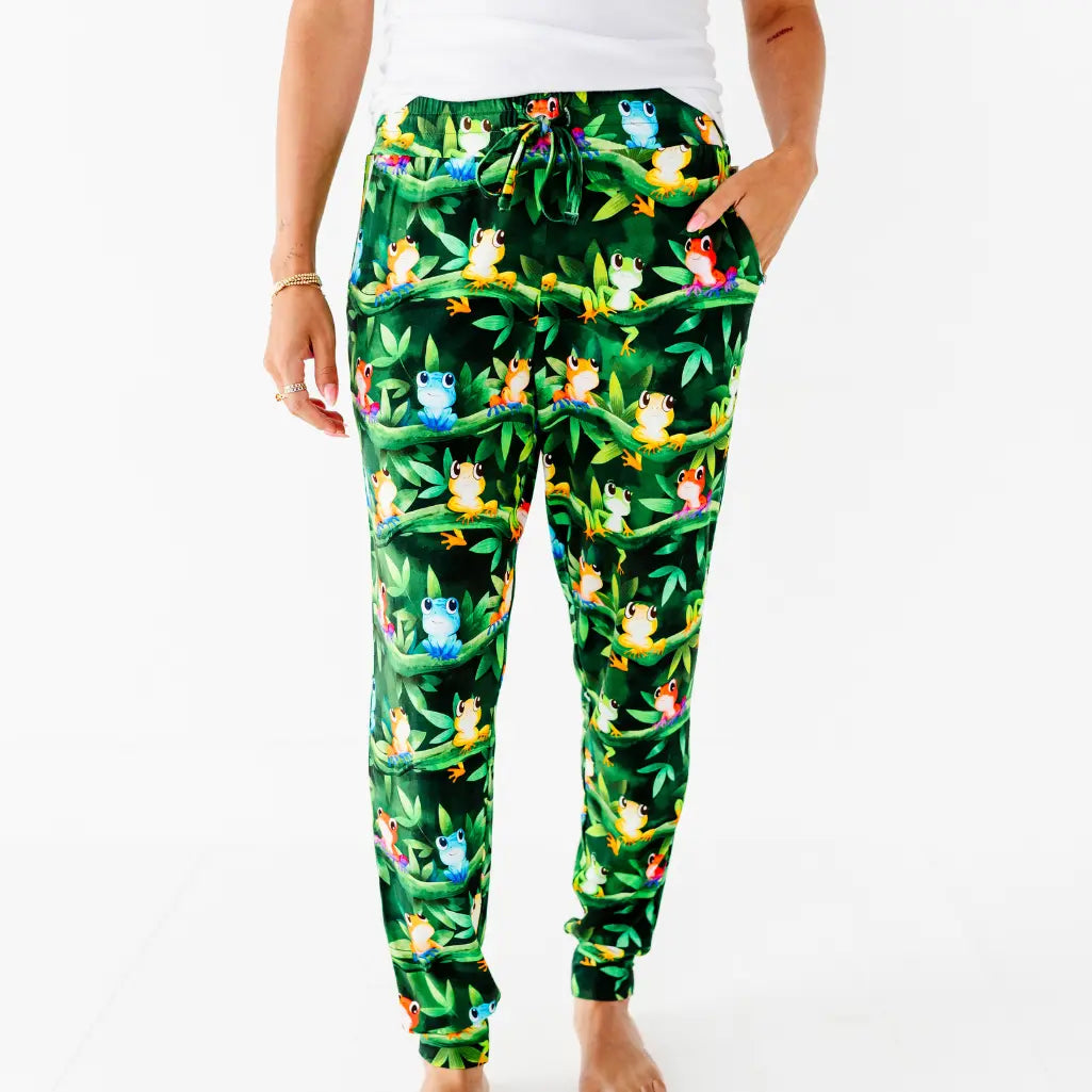 A woman standing with her hand in the pocket of dark green pajama pants covered in colorful tree frogs on leafy vines.