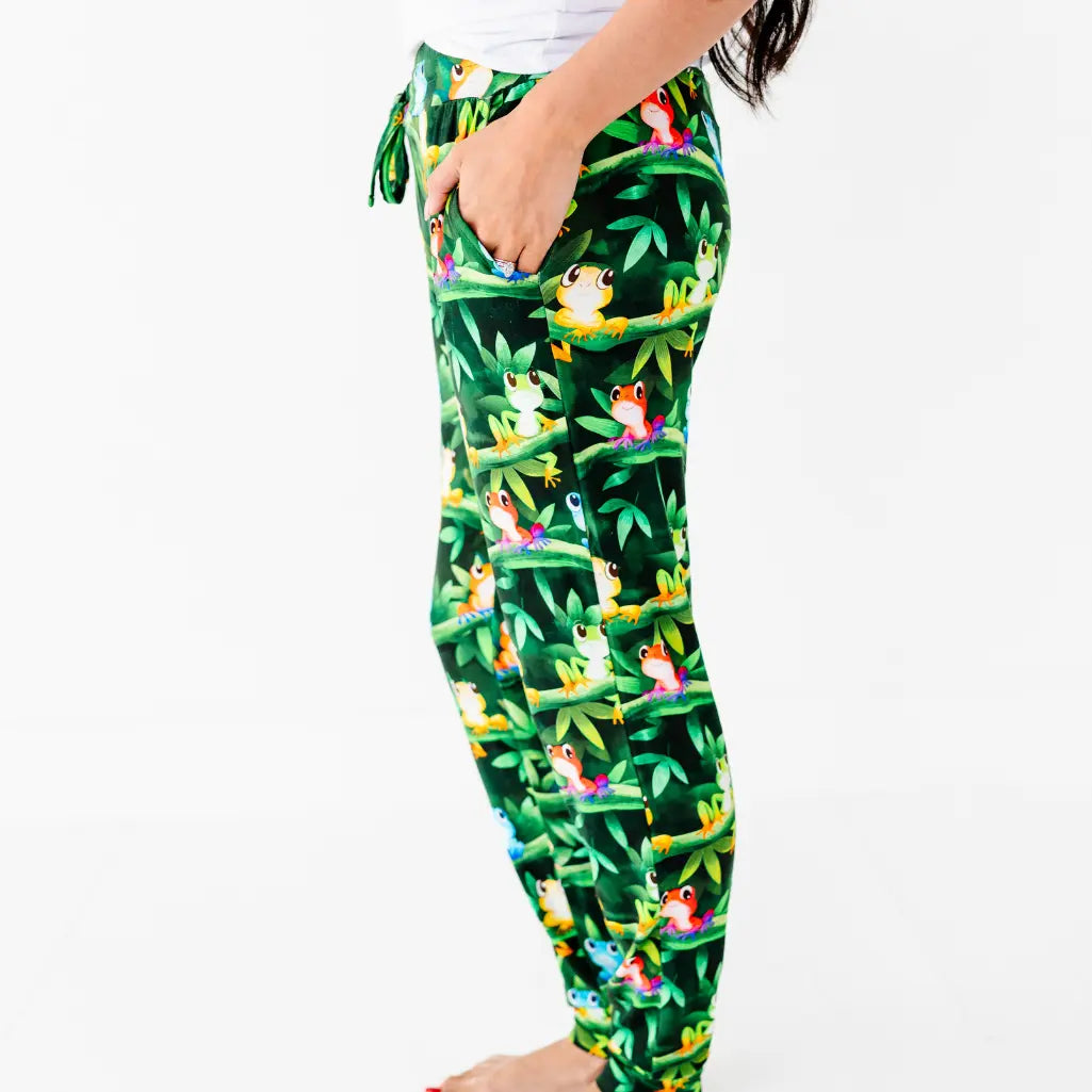 Side view of a woman wearing dark green jogger-style pajama pants with a colorful tree frog and jungle vine print.