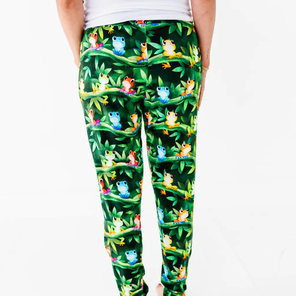 Back view of a woman wearing dark green pajama pants with a colorful tree frog and jungle leaf print.