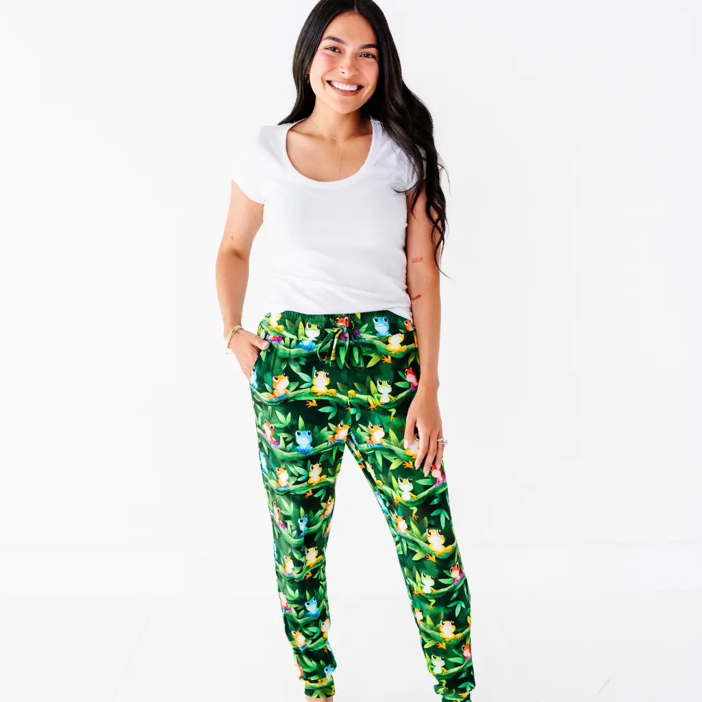 A woman in a white tee smiling with her hand in the pocket of dark green pajama pants with a colorful tree frog print.