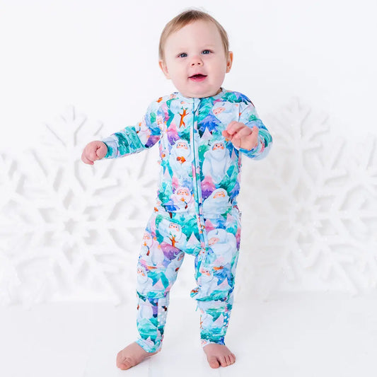 A baby smiling at the camera, standing barefoot with arms out in a zip up pajama with a print of friendly white yetis among teal, blue, and purple pine trees.