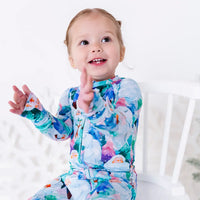 A toddler laughing with hands raised while sitting on a white chair, in a zip up pajama with a print of friendly white yetis among teal, blue, and purple pine trees.