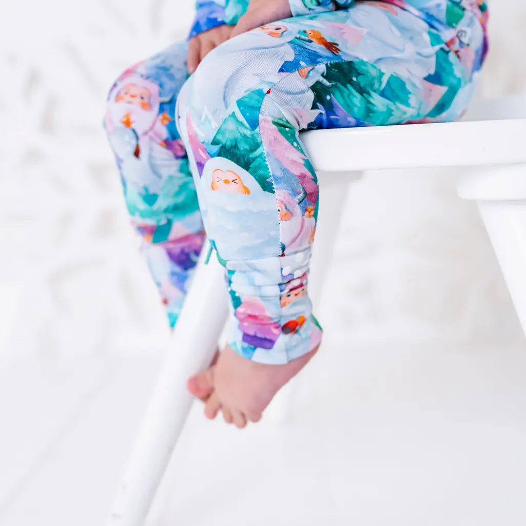 Close up of a toddler's leg and bare foot hanging from a white chair, in a pajama with a print of friendly white yetis among teal, blue, and purple pine trees and a gathered ankle cuff.