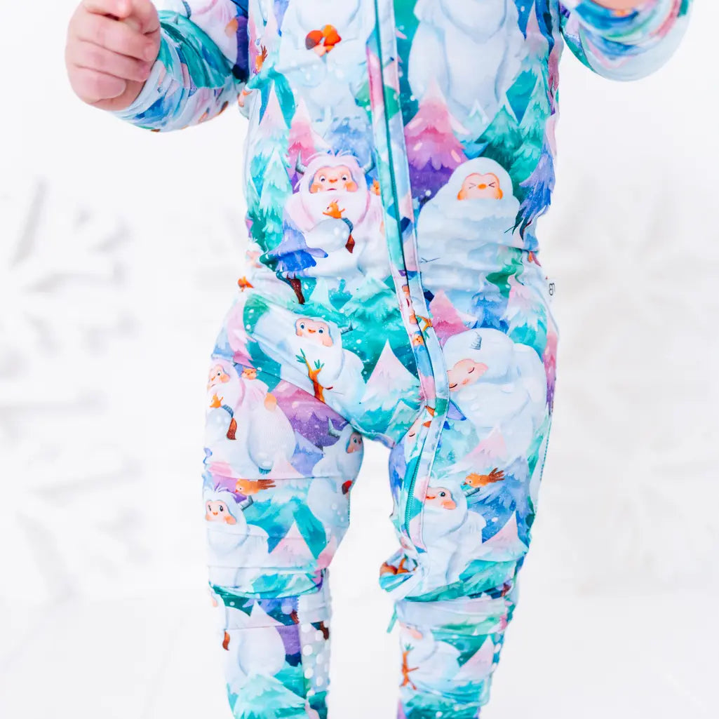 Close up of a baby standing in a zip up pajama, showing the zipper running down the front and into the leg, in a print of friendly white yetis among teal, blue, and purple pine trees.