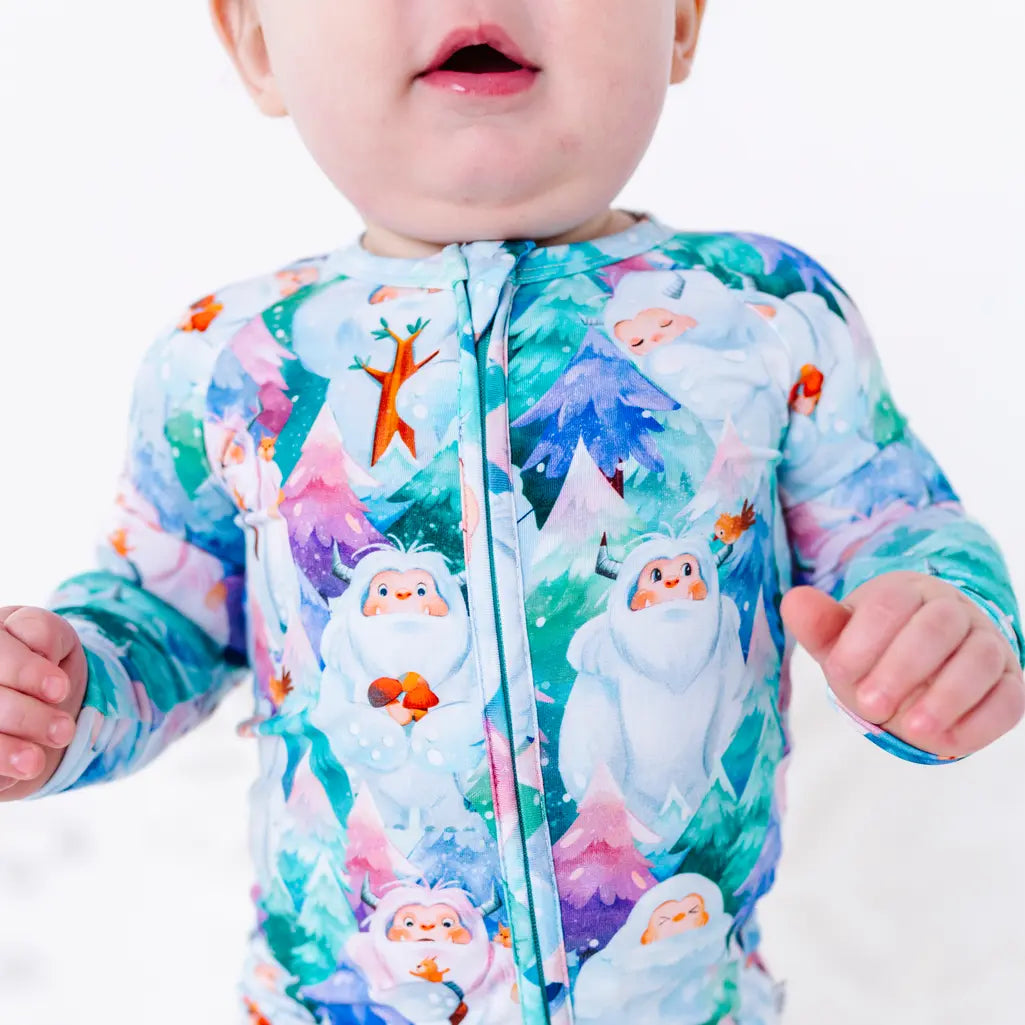 Close up of the front zipper on a baby's zip up pajama with a print of friendly white yetis among teal, blue, and purple pine trees.