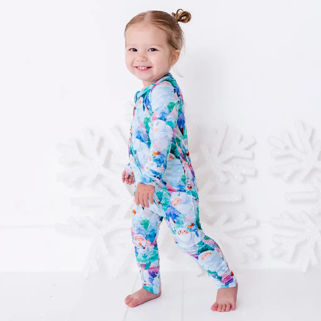A smiling toddler walking barefoot in a zip up pajama with a print of friendly white yetis among teal, blue, and purple pine trees, in front of large white snowflakes.