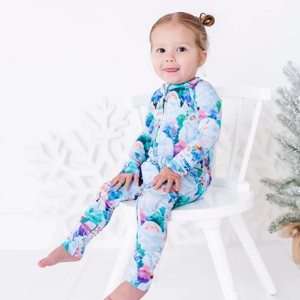A smiling toddler sitting on a white chair with one leg stretched out, in a zip up pajama with a print of friendly white yetis among teal, blue, and purple pine trees.