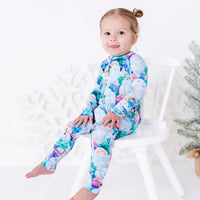 A smiling toddler sitting on a white chair with one leg stretched out, in a zip up pajama with a print of friendly white yetis among teal, blue, and purple pine trees.