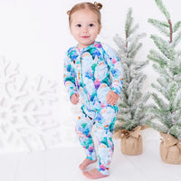 A toddler standing barefoot and looking at the camera, in a zip up pajama with a print of friendly white yetis among teal, blue, and purple pine trees, beside small frosted trees.