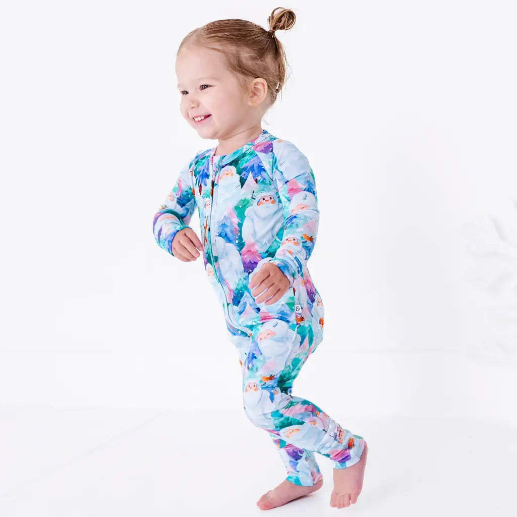 A toddler walking and smiling with arms swinging, in a zip up pajama with a print of friendly white yetis among teal, blue, and purple pine trees.