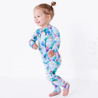 A toddler walking and smiling with arms swinging, in a zip up pajama with a print of friendly white yetis among teal, blue, and purple pine trees.
