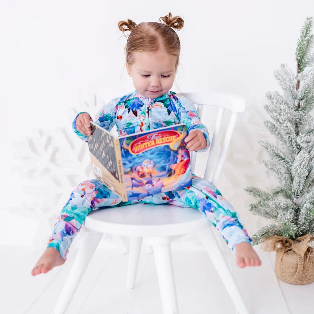 A toddler sitting on a white chair reading a storybook, in a zip up pajama with a print of friendly white yetis among teal, blue, and purple pine trees.