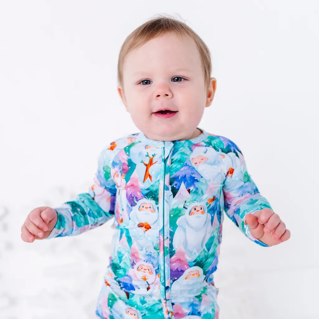 A baby smiling at the camera in a zip up pajama with a print of friendly white yetis among teal, blue, and purple pine trees, shown from the waist up.