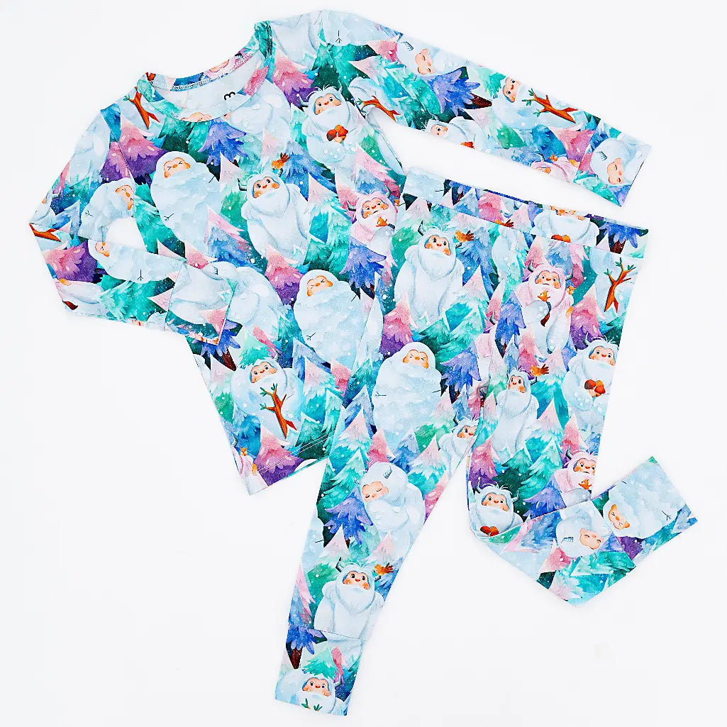 Flat lay of a long sleeve pajama top and matching pants in a print of friendly white yetis among teal, blue, and purple pine trees on a white background.