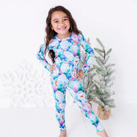 A smiling child with hands on hips in a long sleeve pajama set with a print of friendly white yetis among teal, blue, and purple pine trees, standing beside small frosted trees.