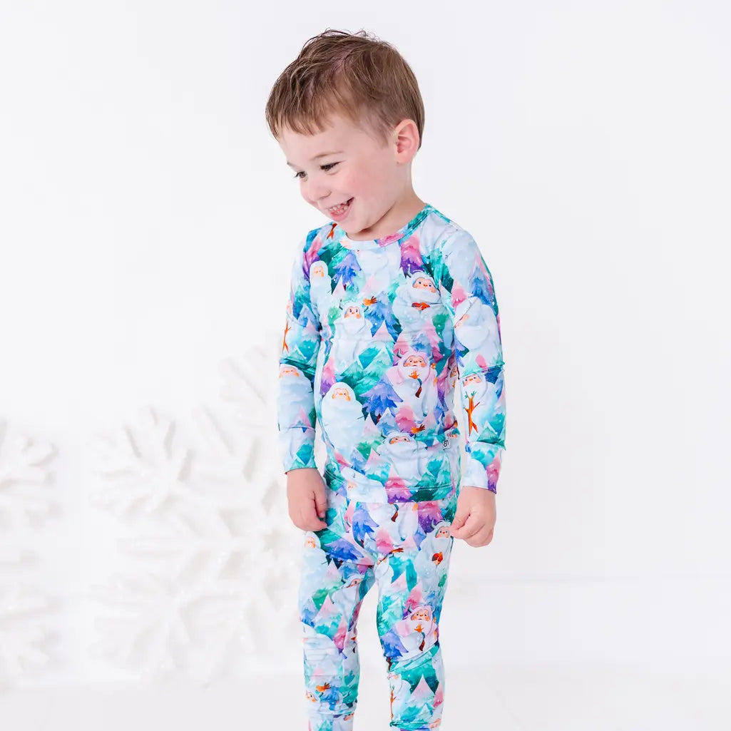 A toddler laughing and looking down while turned to the side, wearing a long sleeve pajama set with a print of friendly white yetis among teal, blue, and purple pine trees.