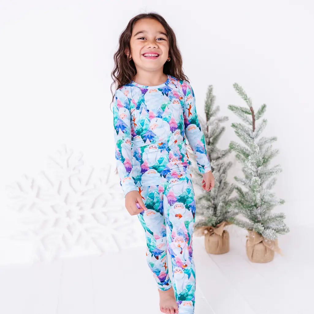 A smiling child walking toward the camera in a long sleeve pajama set with a print of friendly white yetis among teal, blue, and purple pine trees, with small frosted trees behind.