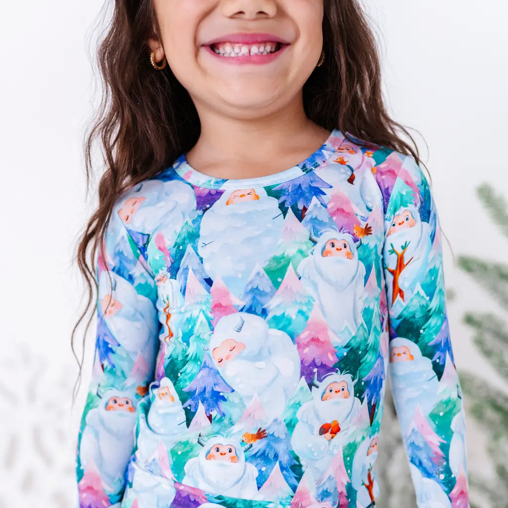 Close up of a child's big smile above a long sleeve pajama top with a print of friendly white yetis among teal, blue, and purple pine trees.