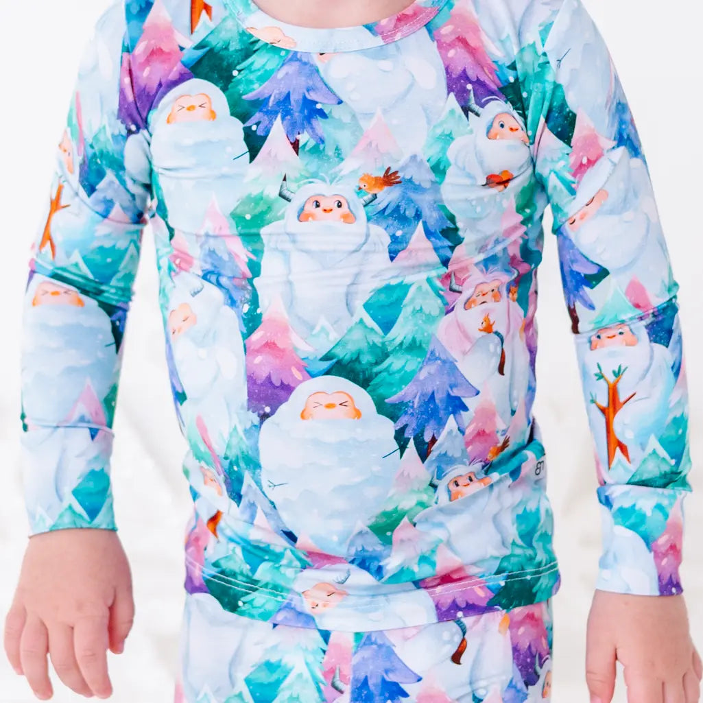 Close up of a toddler standing with arms relaxed at the sides in a long sleeve pajama top with a print of friendly white yetis among teal, blue, and purple pine trees.