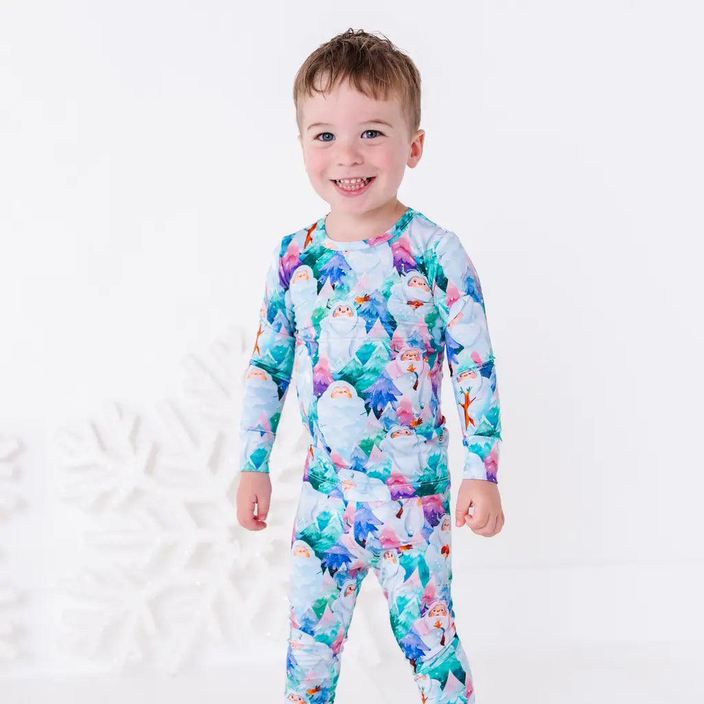 A smiling toddler in a long sleeve pajama set with a print of friendly white yetis among teal, blue, and purple pine trees, standing in front of a large white snowflake.
