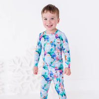 A smiling toddler in a long sleeve pajama set with a print of friendly white yetis among teal, blue, and purple pine trees, standing in front of a large white snowflake.