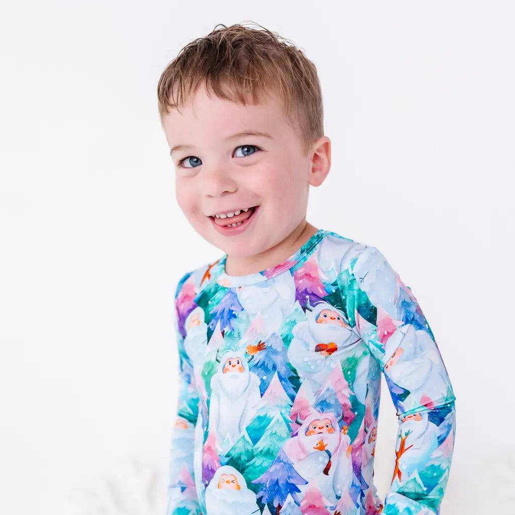 A toddler smiling with head tilted, wearing a long sleeve pajama top with a print of friendly white yetis among teal, blue, and purple pine trees.