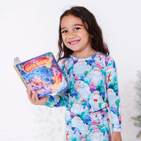 A smiling child holding a storybook in a long sleeve pajama set with a print of friendly white yetis among teal, blue, and purple pine trees.